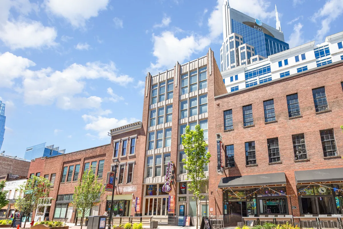 This exterior scene captures a lively downtown Nashville street featuring a historic brick building juxtaposed with a sleek modern skyscraper. Green trees and an inviting outdoor seating area create a dynamic mix of work, dining, and exploration possibilities just steps from the property.