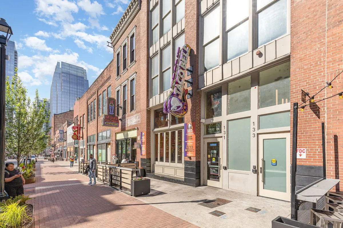A bustling streetscape featuring a Taco Bell sign among various storefronts, people passing by, and lively outdoor seating. The scene captures the energetic vibe of Nashville’s social spaces, with groups dining and socializing.