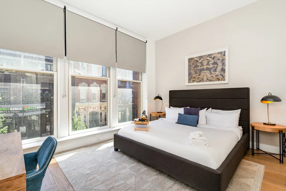 A serene bedroom featuring a neatly made bed with an abundance of pillows, large windows that let in natural light, tasteful wall art, and nightstands with lamps. The room suggests both relaxation and the presence of a small desk or workstation for productivity.
