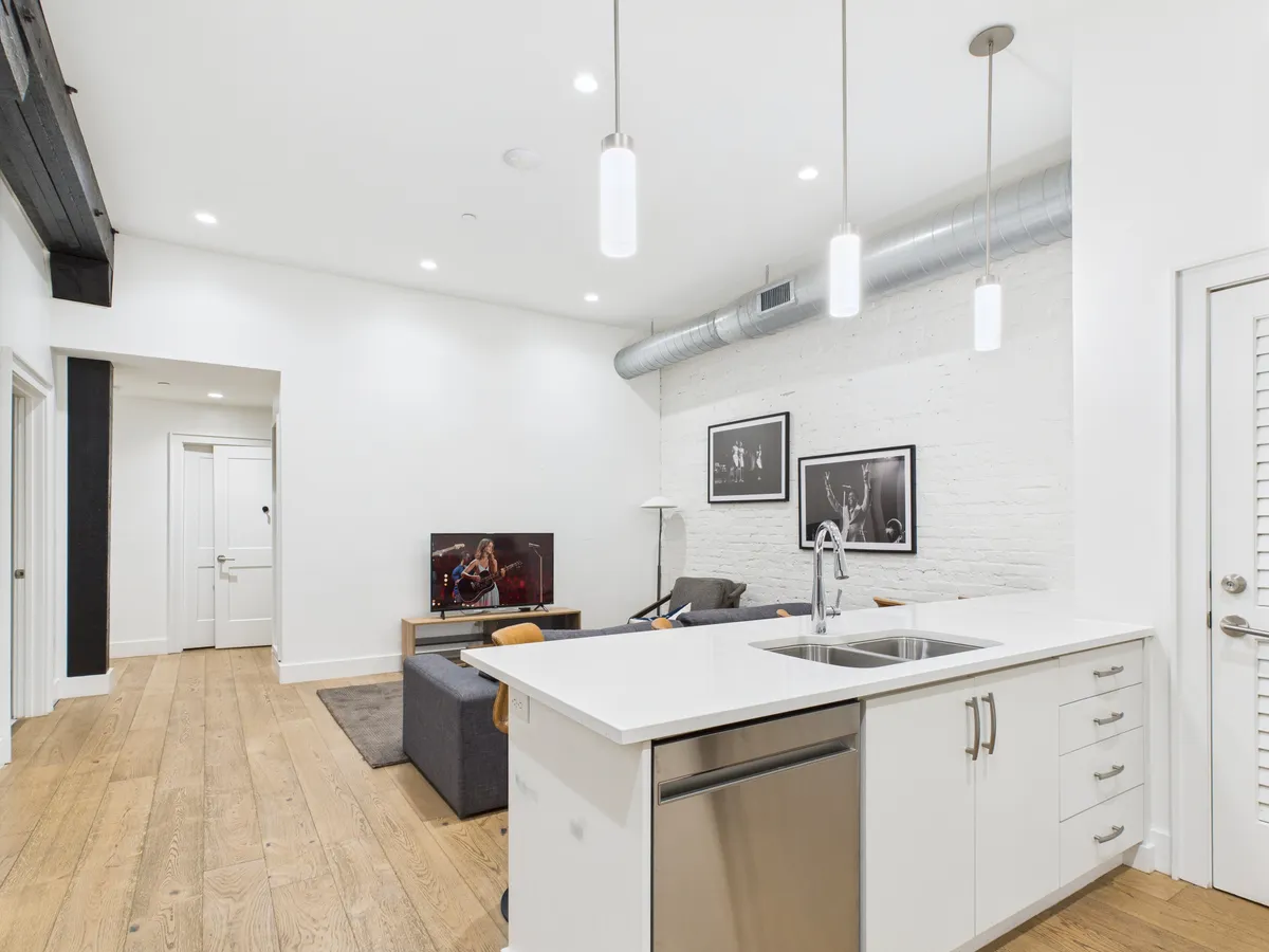 A contemporary open-concept living space featuring a plush sofa, kitchen island, television, and eye-catching artwork. The area is bathed in natural light, with high ceilings and a seamless flow between the living and kitchen zones, emphasizing communal living and entertainment.