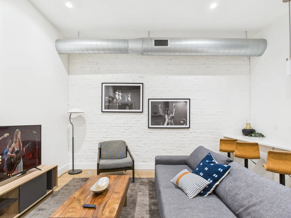 A modern living room setup with a gray sofa adorned with accent pillows, a wall-mounted TV displaying a live performance, a wooden coffee table, and stylish black-and-white artwork. The space feels inviting and is clearly designed for relaxation, conversation, and entertainment.
