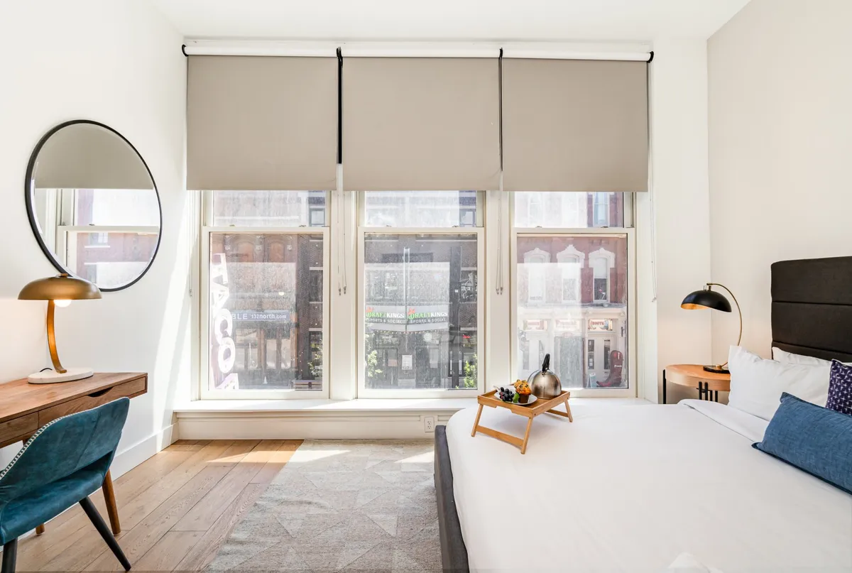 A contemporary bedroom highlighted by large windows with sweeping city views, a comfortable bed adorned with decorative pillows, a stylish mirror, a sleek desk and chair, and a tray of fruit, suggesting a blend of relaxation and work possibilities.